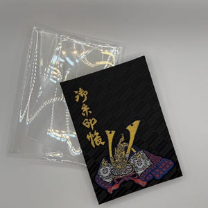 Puede incluir: Una tarjeta rectangular negra con texto japonés dorado y una ilustración detallada de un casco de samurái en rojo, azul y dorado. La tarjeta está junto a una funda de plástico transparente.