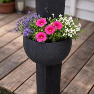 Può includere: Una fioriera nera, semi-sferica, piena di fiori vibranti, tra cui petunie rosa brillante, lavanda viola e alyssum bianco. La fioriera è montata su un palo di legno nero, creando un contrasto sorprendente con il decking in legno naturale.