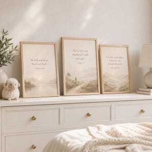 May include: Three framed wall art prints with light wood frames, each featuring a beige landscape illustration and religious text. The left print reads Be still and know that I am God, the center reads The Lord is my shepherd, and the right reads For I know the plans I have for you.