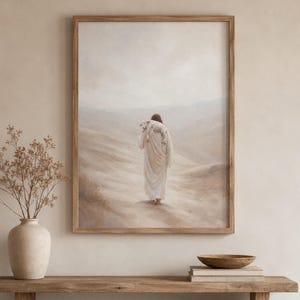 May include: A framed wall art print depicting a figure in a long, flowing cream-colored robe walking away through a hazy, desert-like landscape while carrying a small white lamb draped over their shoulders, rendered in soft, muted earth tones.