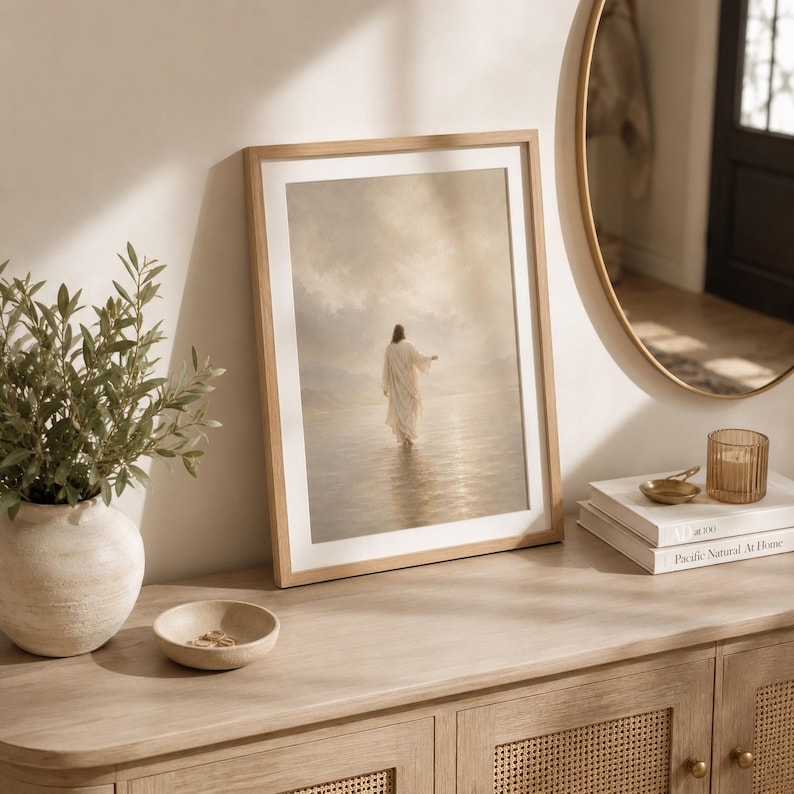 May include: A framed vertical wall art print featuring a figure in a long white robe walking across a calm, reflective body of water under a cloudy, soft-toned sky. The artwork is encased in a light-colored wooden frame with a white mat border.