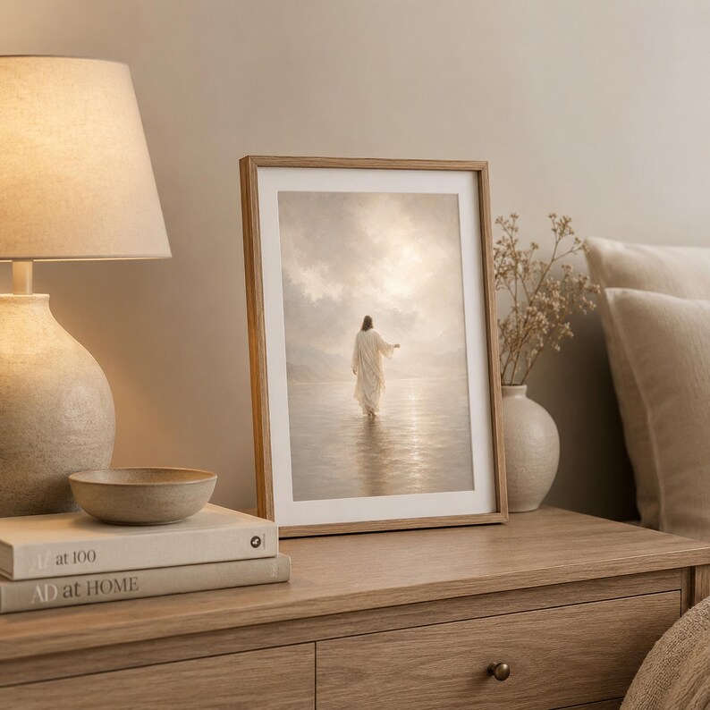 May include: A framed vertical print depicting a figure in a long white robe walking across a calm, reflective body of water toward a bright, cloudy horizon. The artwork is displayed in a light wood frame, positioned on a wooden surface.