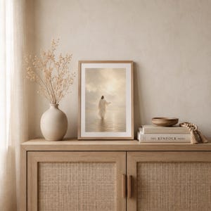 May include: A framed vertical wall art print depicting a figure in a white robe walking across a calm body of water toward a bright, cloudy horizon. The artwork is displayed in a light wood frame, positioned next to a beige vase with dried stems.