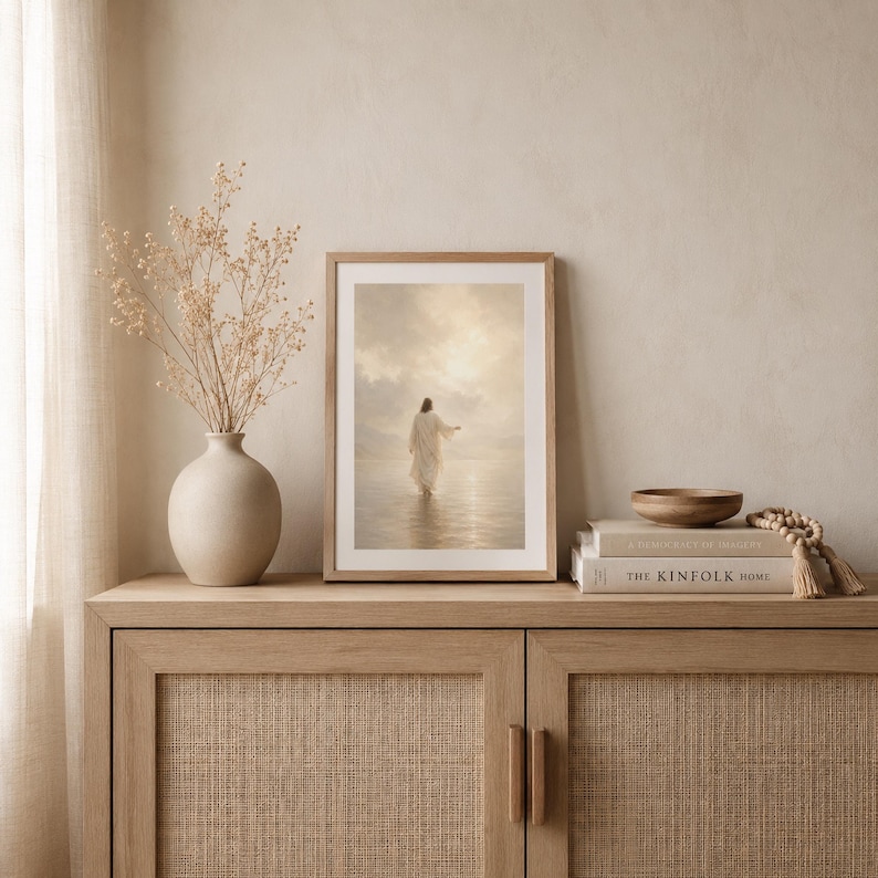 May include: A framed vertical wall art print depicting a figure in a white robe walking across a calm body of water toward a bright, cloudy horizon. The artwork is displayed in a light wood frame, positioned next to a beige vase with dried stems.