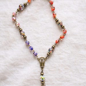 Puede incluir: Un rosario con cuentas de vidrio de colores en rojo, naranja, azul y diseños florales. Las cuentas están ensartadas en una cadena de color bronce, con un colgante que representa una figura religiosa.
