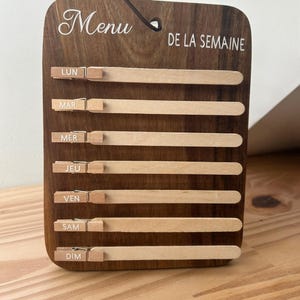 Peut inclure: Tableau de menu en bois avec l'inscription "Menu DE LA SEMAINE" en écriture blanche. Il comporte sept lattes en bois avec de petites pinces, chacune étiquetée avec un jour de la semaine de LUN à DIM. Une corde marron foncé est attachée pour l'accrocher.