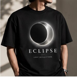 May include: A black short-sleeved t-shirt featuring a graphic of a glowing solar eclipse. Below the eclipse, the word ECLIPSE is printed in white capital letters, followed by the text LIGHT REVEALS FORM in smaller white capital letters.