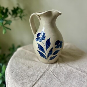 May include: A beige ceramic pitcher featuring a hand-painted blue floral design on the front. The vessel has a curved handle, a rounded body, and a wide, scalloped pouring spout. The surface of the pottery shows a smooth, matte finish.