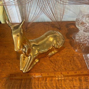 May include: A gold-colored ceramic deer figurine in a reclining position, positioned next to a wooden ruler that shows the object is approximately 11 inches in length. The deer has a smooth, reflective surface and is depicted with its legs tucked.