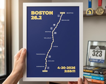 May include: A framed print with a navy blue background displays the text "BOSTON 26.2" in yellow. A yellow line traces a marathon route with place names. The date "4-20-2026" and time "2:55:11" are shown. The print is held by hands.