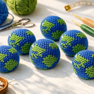 May include: A collection of handmade crochet Earth balls in blue and green, depicting continents. The image also shows blue and green yarn, scissors, crochet hooks, and a measuring tape. A small wooden bowl with pins is also visible.
