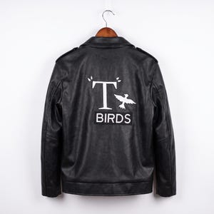 Può includere: Una giacca nera con colletto e maniche lunghe, caratterizzata da una grafica bianca sul retro. Il design include una grande lettera T, la sagoma di un uccello e la parola BIRDS stampata in bianco all'interno di un riquadro rettangolare.