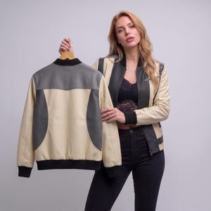 May include: A colour-blocked bomber jacket featuring cream and dark grey panels, a black ribbed collar, cuffs, and hem. The jacket has a front zip closure and long sleeves. The design incorporates a mix of neutral tones with a structured fit.