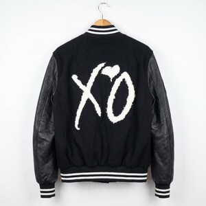 May include: A black varsity jacket featuring long black sleeves and a white embroidered XO logo with a heart on the back. The jacket includes a ribbed collar, cuffs, and hem with white stripes, and is displayed on a wooden hanger.