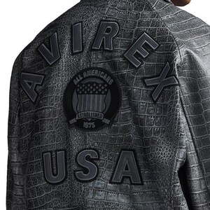 May include: A charcoal grey jacket with a textured crocodile skin pattern. The back features the black embroidered text AVIREX in an arc, the letters USA below, and a circular patch in the center with the text ALL AMERICANS ESTABLISHED 1975.
