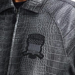 May include: A charcoal gray jacket with a textured crocodile skin pattern. The jacket features a front zipper closure and a black embroidered patch on the chest displaying the text AVIREX, ESTABLISHED, and 1975, along with a shield and star graphic.