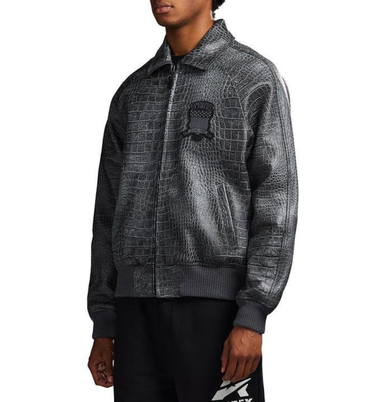 May include: A charcoal gray bomber jacket featuring a textured crocodile skin pattern throughout. The jacket includes a front zipper closure, a pointed collar, ribbed cuffs and hem, and a dark circular patch on the left chest with text.