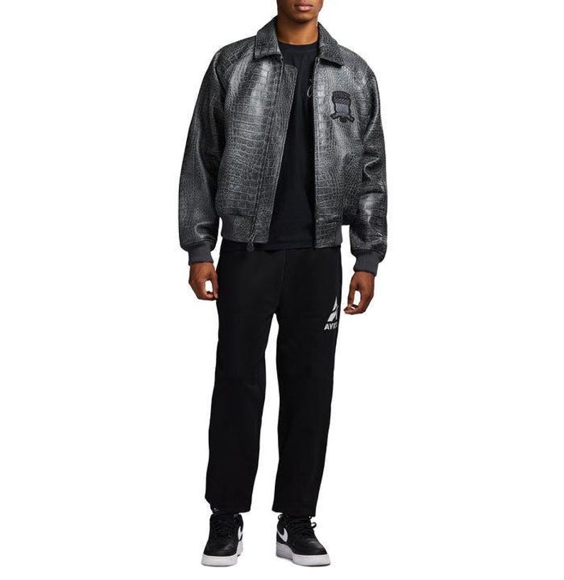 May include: A charcoal gray bomber jacket with a textured crocodile skin pattern, featuring a pointed collar, ribbed cuffs, and a dark embroidered patch on the chest. It is worn over a plain black t-shirt, black sweatpants, and black and white sneakers.