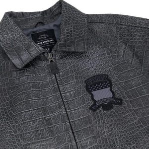 May include: A charcoal gray jacket with a textured crocodile skin pattern. The jacket features a front zipper closure, a collar, and a black patch on the chest with the text AVIREX ESTABLISHED. An interior label reads AUTHENTIC 1975 AVIREX.