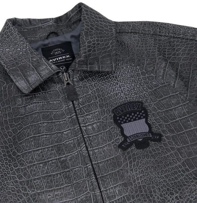 May include: A charcoal gray jacket with a textured crocodile skin pattern. The jacket features a front zipper closure, a collar, and a black patch on the chest with the text AVIREX ESTABLISHED. An interior label reads AUTHENTIC 1975 AVIREX.