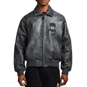 May include: A charcoal grey bomber jacket featuring a textured crocodile skin pattern, a classic collar, and a full-length front zipper. A black embroidered patch with a shield design and text is positioned on the upper left chest area.