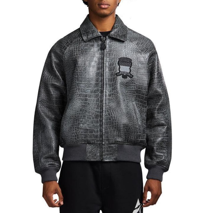 May include: A charcoal grey bomber jacket featuring a textured crocodile skin pattern, a classic collar, and a full-length front zipper. A black embroidered patch with a shield design and text is positioned on the upper left chest area.