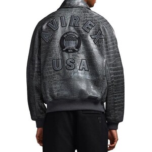 May include: A grey leather bomber jacket with a textured crocodile skin pattern. The back features the text AVIREX in large letters, a circular patch with the text ALL AMERICANS ESTABLISHED 1975, and the text USA below it. It has a ribbed hem.