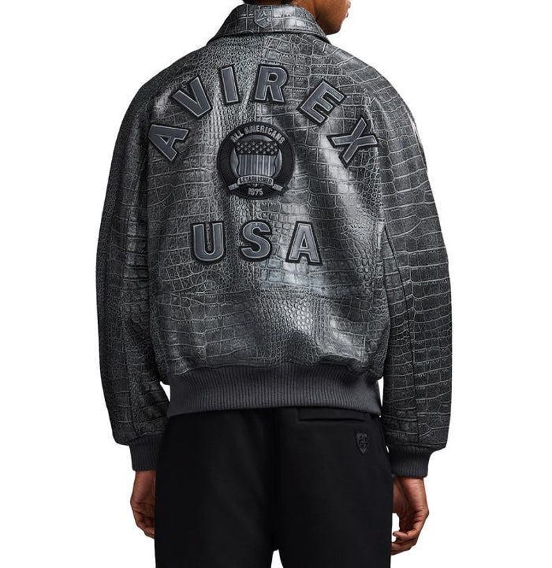 May include: A grey leather bomber jacket with a textured crocodile skin pattern. The back features the text AVIREX in large letters, a circular patch with the text ALL AMERICANS ESTABLISHED 1975, and the text USA below it. It has a ribbed hem.