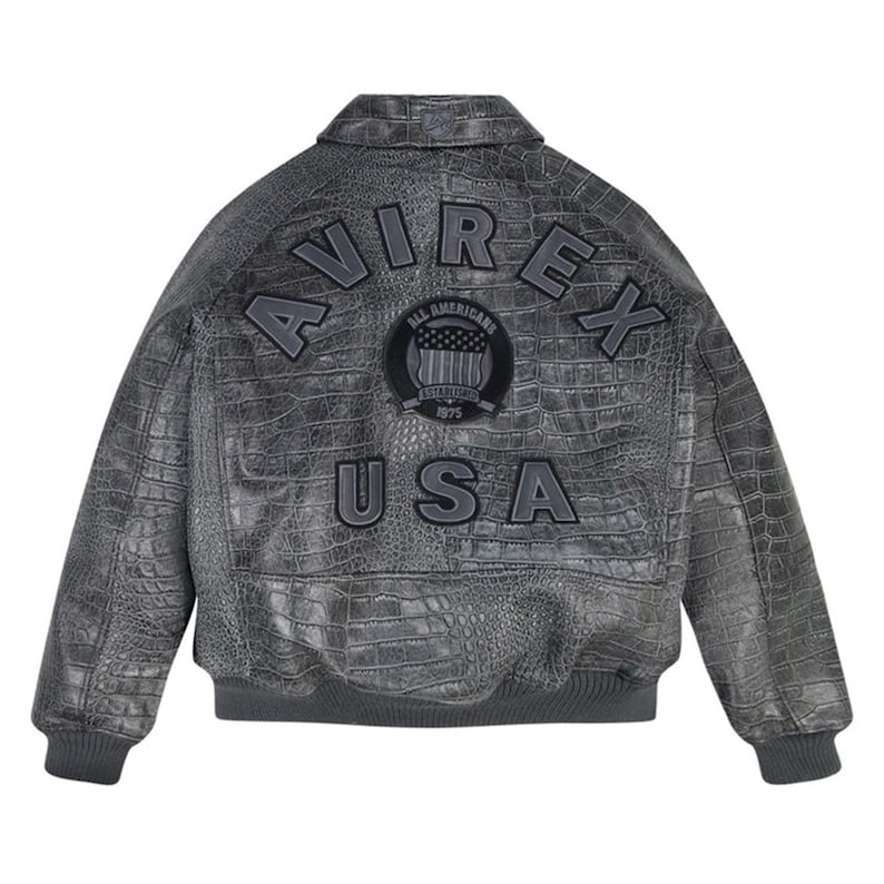 May include: A charcoal grey leather jacket with a textured crocodile skin pattern. The back features the text AVIREX in an arch, a circular patch with the text ALL AMERICANS ESTABLISHED 1975, and the text USA below. It has a ribbed hem and cuffs.