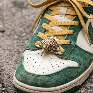 May include: Close-up of a green and white sneaker with gold shoelaces and a gold eagle head lace lock. The shoe has a green suede overlay and a white leather base with a perforated toe box. The laces are a mustard yellow.