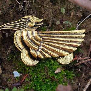 May include: A gold-coloured metal bird pin with detailed wings and a long beak. The pin is set against a backdrop of soil, moss, and small twigs, creating a natural contrast. The bird's design is intricate, with visible feather detailing.