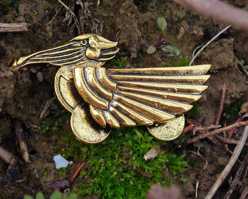 May include: A gold-coloured metal bird pin with detailed wings and a long beak. The pin is set against a backdrop of soil, moss, and small twigs, creating a natural contrast. The bird's design is intricate, with visible feather detailing.