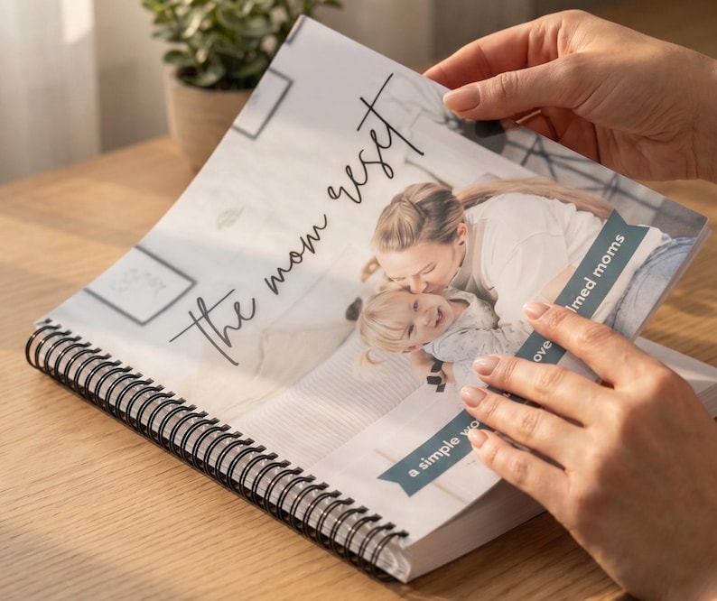 May include: A spiral-bound journal titled "the mom reset" with a photograph of a mother and child. The journal is open, revealing the image and text. The cover features a soft, neutral color palette.