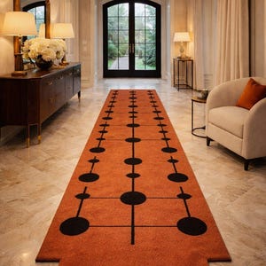 May include: An orange runner rug with a geometric black pattern in a hallway. The rug features a series of connected circles and lines. The hallway has a marble floor, a dark wood cabinet, and a large mirror.