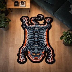 May include: A decorative rug in the shape of a tiger, featuring a black outline with orange and blue details. The rug is placed on a light wood floor, with a sofa and plants in the background.