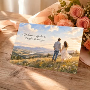 May include: A rectangular greeting card featuring a scenic landscape with a couple standing in a wildflower field. The text reads, Wherever life leads, I'm glad it's with you. The background shows mountains, a river, and a sunset sky.