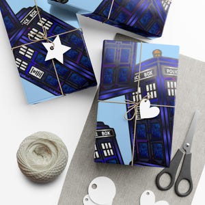Puede incluir: Cajas de regalo envueltas con un diseño de Tardis azul y morado. Las cajas están atadas con hilo y adornadas con etiquetas blancas en forma de estrella y corazón. También hay una madeja de hilo, tijeras y etiquetas en blanco.