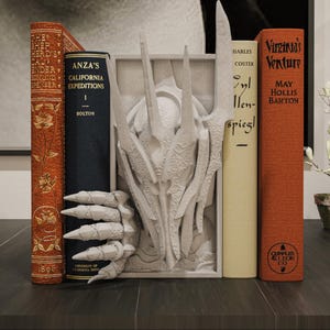 May include: A decorative bookend with a white dragon design, holding several vintage books. The books are in various colours, including orange, cream, and navy blue, with gold lettering and floral patterns. The bookend is on a dark wooden surface.