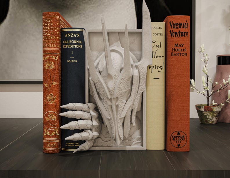 May include: A decorative bookend with a white dragon design, holding several vintage books. The books are in various colours, including orange, cream, and navy blue, with gold lettering and floral patterns. The bookend is on a dark wooden surface.