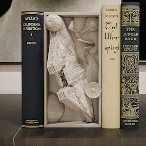 Könnte beinhalten: Eine weiße, rechteckige Buchstütze mit einem detaillierten, skulpturalen Modell eines Raumschiffs und einer kugelförmigen Raumstation. Die Buchstütze steht zwischen zwei Büchern, eines mit dunkelblauem und eines mit cremefarbenem Rücken.