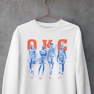 Op de afbeelding: Witte sweatshirt met een blauw en oranje afbeelding van vier basketballers. Het woord "OKC" is in oranje boven de spelers gedrukt. Elke speler heeft een handtekening eronder. De sweatshirt hangt aan een metalen hanger.