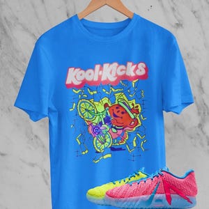 Könnte beinhalten: Ein leuchtend blaues Kurzarm-T-Shirt mit einer Grafik eines roten krugförmigen Charakters auf einem Fahrrad und dem Schriftzug Kool-Kicks in weißen Buchstaben. Darunter befinden sich zwei Sneaker mit neongelbem und pinkfarbenem Obermaterial.
