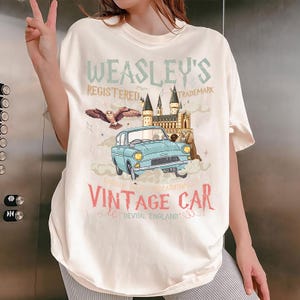May include: Off-white t-shirt with a light blue vintage car graphic, text reading "Weasley's Registered Trademark" and "Vintage Car, Devon, England." The design incorporates an owl, a castle, and clouds.