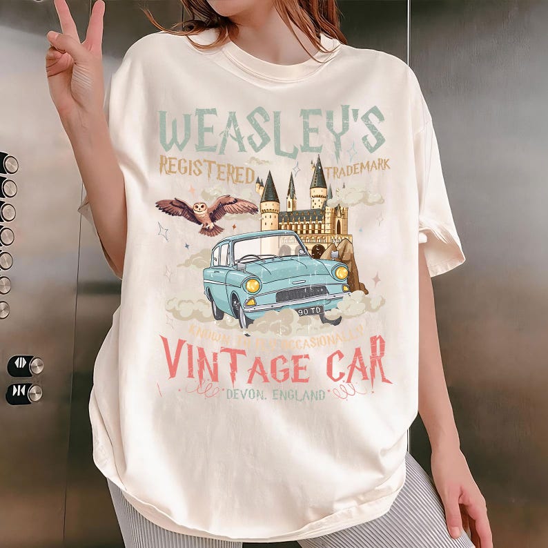 May include: Off-white t-shirt with a light blue vintage car graphic, text reading "Weasley's Registered Trademark" and "Vintage Car, Devon, England." The design incorporates an owl, a castle, and clouds.