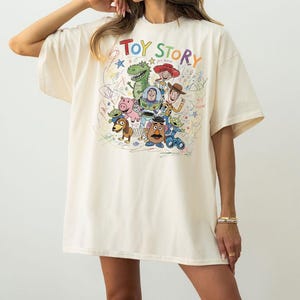 May include: An oversized cream-colored t-shirt featuring a colorful graphic print of Toy Story characters including Woody, Buzz Lightyear, Jessie, Rex, Slinky Dog, Mr. Potato Head, Hamm, and Aliens, surrounded by multi-colored scribbles and stars.