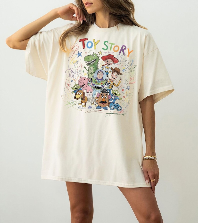 May include: An oversized cream-colored t-shirt featuring a colorful graphic print of Toy Story characters including Woody, Buzz Lightyear, Jessie, Rex, Slinky Dog, Mr. Potato Head, Hamm, and Aliens, surrounded by multi-colored scribbles and stars.