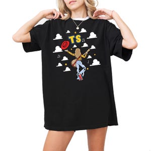 May include: A black t-shirt featuring a graphic of a person with long blonde hair wearing a yellow plaid shirt and blue cow-print bell-bottom jeans. The person holds a red hat and a guitar. The text TS is above, surrounded by white clouds and stars.