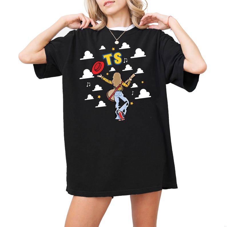 May include: A black t-shirt featuring a graphic of a person with long blonde hair wearing a yellow plaid shirt and blue cow-print bell-bottom jeans. The person holds a red hat and a guitar. The text TS is above, surrounded by white clouds and stars.