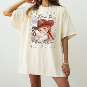 May include: An oversized cream-coloured t-shirt featuring a graphic print of a cartoon cowgirl with red hair in a braid, wearing a brown cowboy hat and cow-print glove. The text reads EST 2026, I Knew It, and I Knew You, framed by stars and horseshoes.