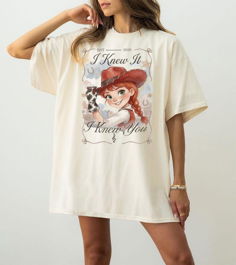 May include: An oversized cream-coloured t-shirt featuring a graphic print of a cartoon cowgirl with red hair in a braid, wearing a brown cowboy hat and cow-print glove. The text reads EST 2026, I Knew It, and I Knew You, framed by stars and horseshoes.